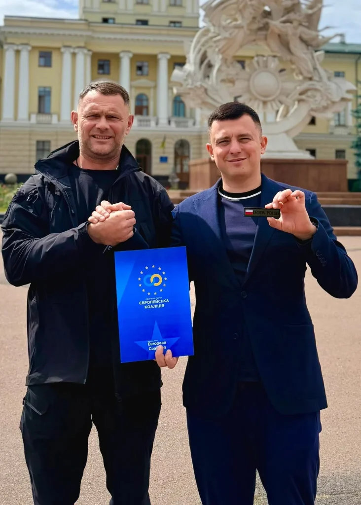 Two men holding a folder with the «Європейська Коаліція» emblem and a Legion Polski patch